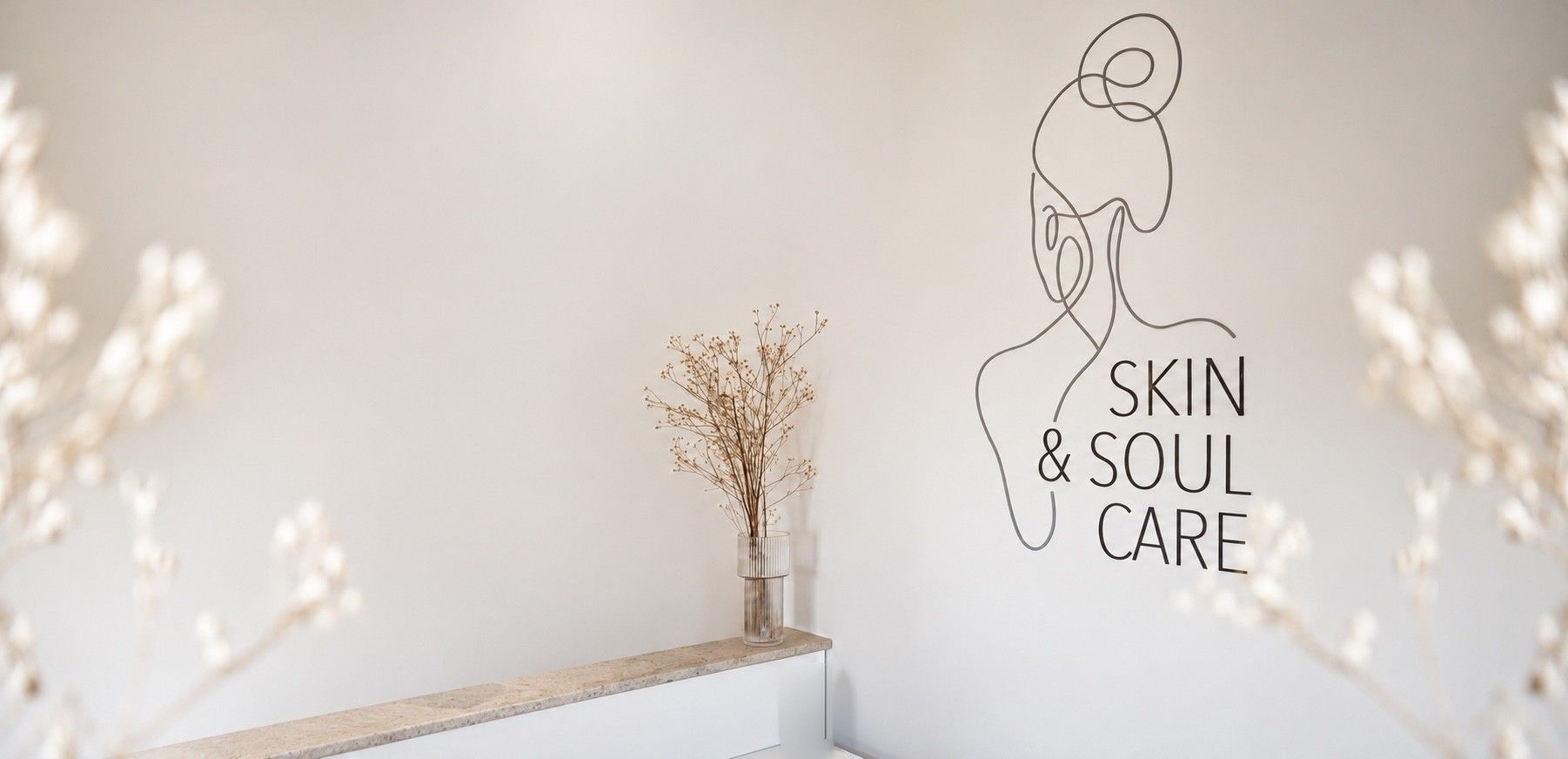 Heller Empfangsraum mit dem Logo an der Wand, davor eine Holzbank mit Trockenblumen