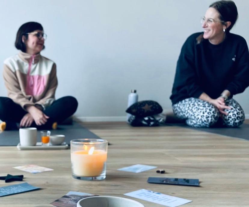 Zwei jüngere Frauen sitzen auf Yoga-Matten und unterhalten sich lachend